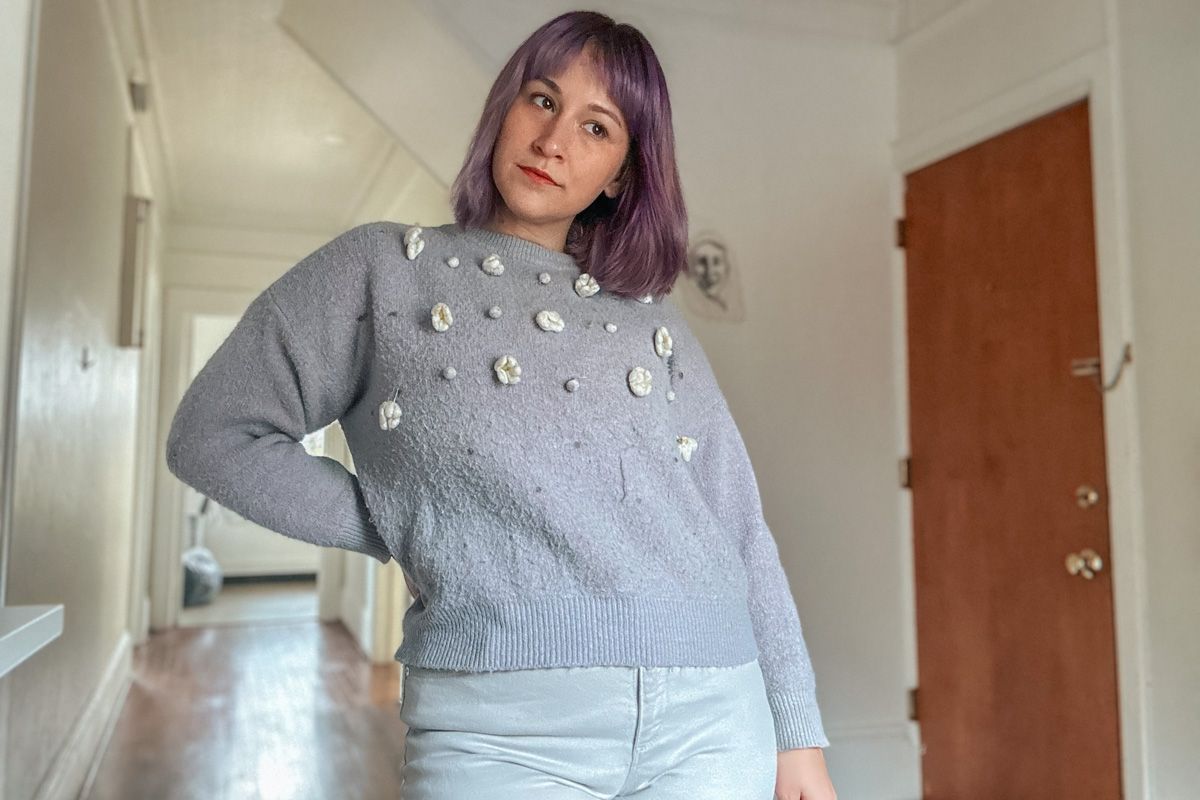 A woman with purple hair wears a light purple sweater with white flowers and her head to the side with a hand on her hip, a sparse interior in soft focus behind her.
