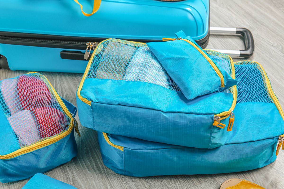 A close-up view of a set of three blue packing cubes with a mesh window, sitting on the floor in front of a matching blue, hard-shell suitcase.