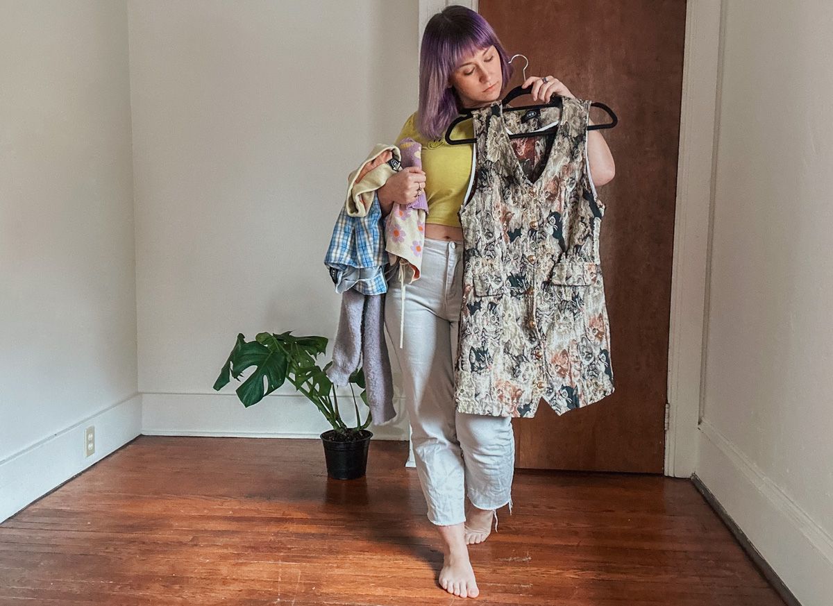 A sparse apartment interior with a woman with purple hair wearing a yellow shirt holding a stack of clothing and a brown dress on a hanger up to her body in preparation to write a Shop Cider review.