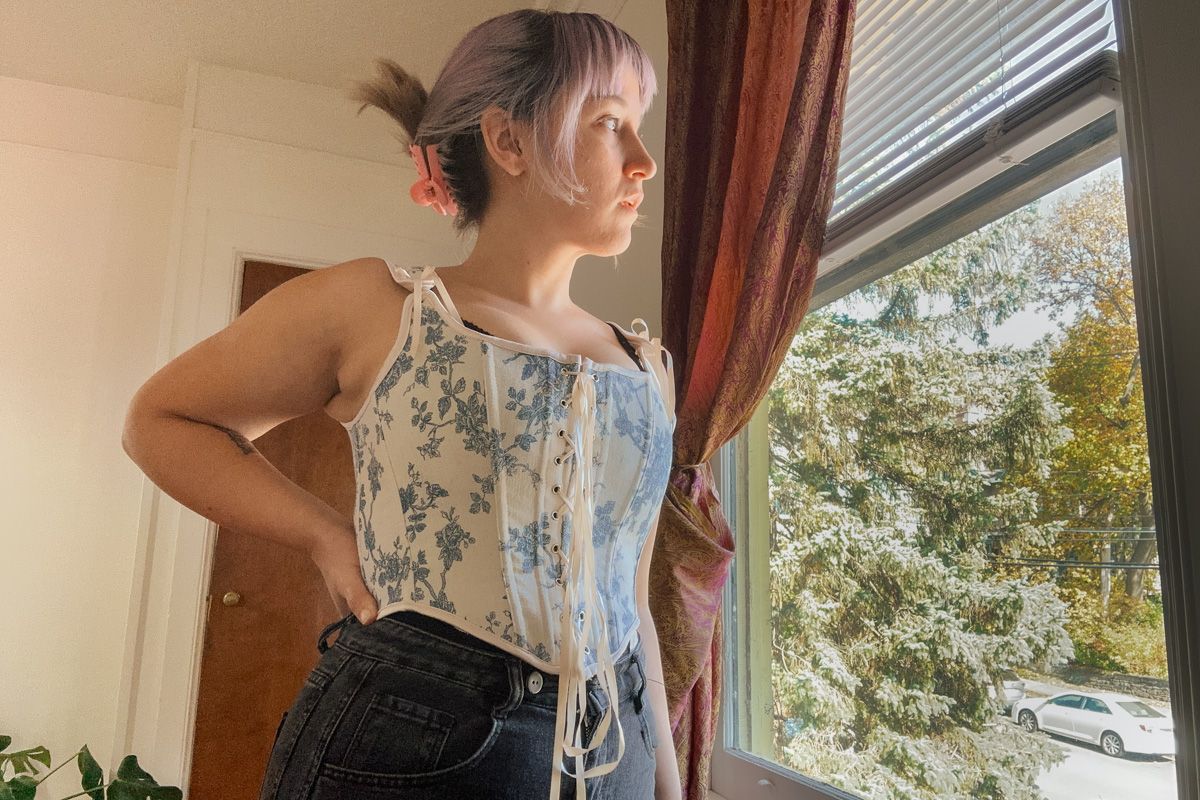 A purple-haired woman wearing a blue-and-white toile corset top stands with a hand on her hip in a sparse interior space and looks out the window at leafy branches.