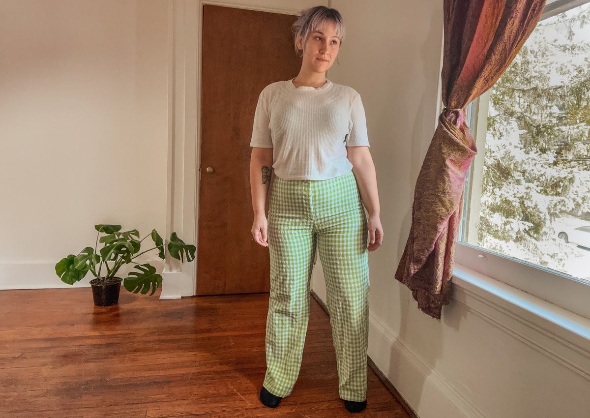 A purple-haired woman wearing a white t-shirt and a pair of green and white checkered pants stands in a sparse interior space beside a window where leafy branches are visible outside.