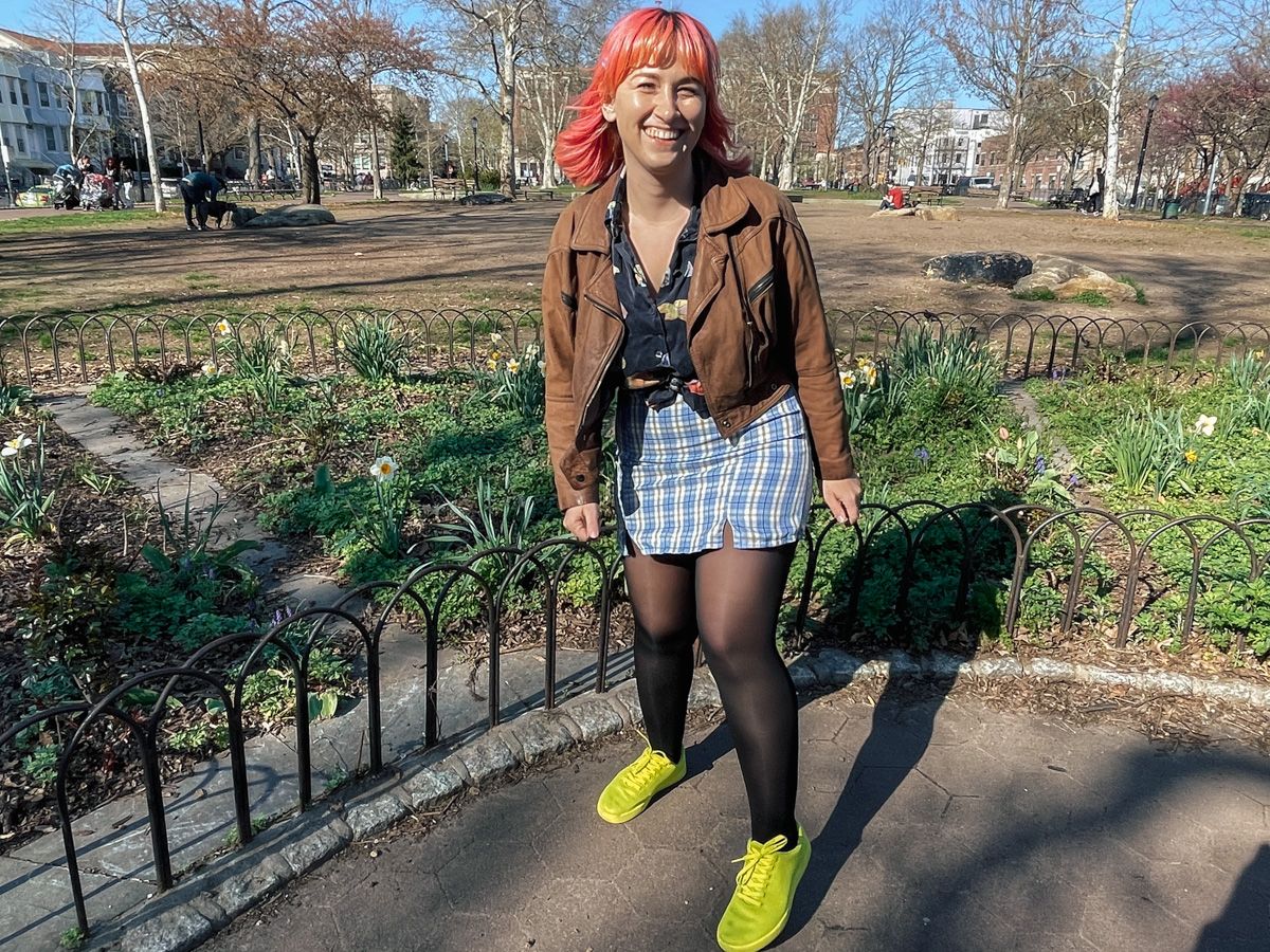 A woman with pink hair in a brown leather jacket, a blue plaid miniskirt, and neon green shoes laughs towards the camera in a city park.