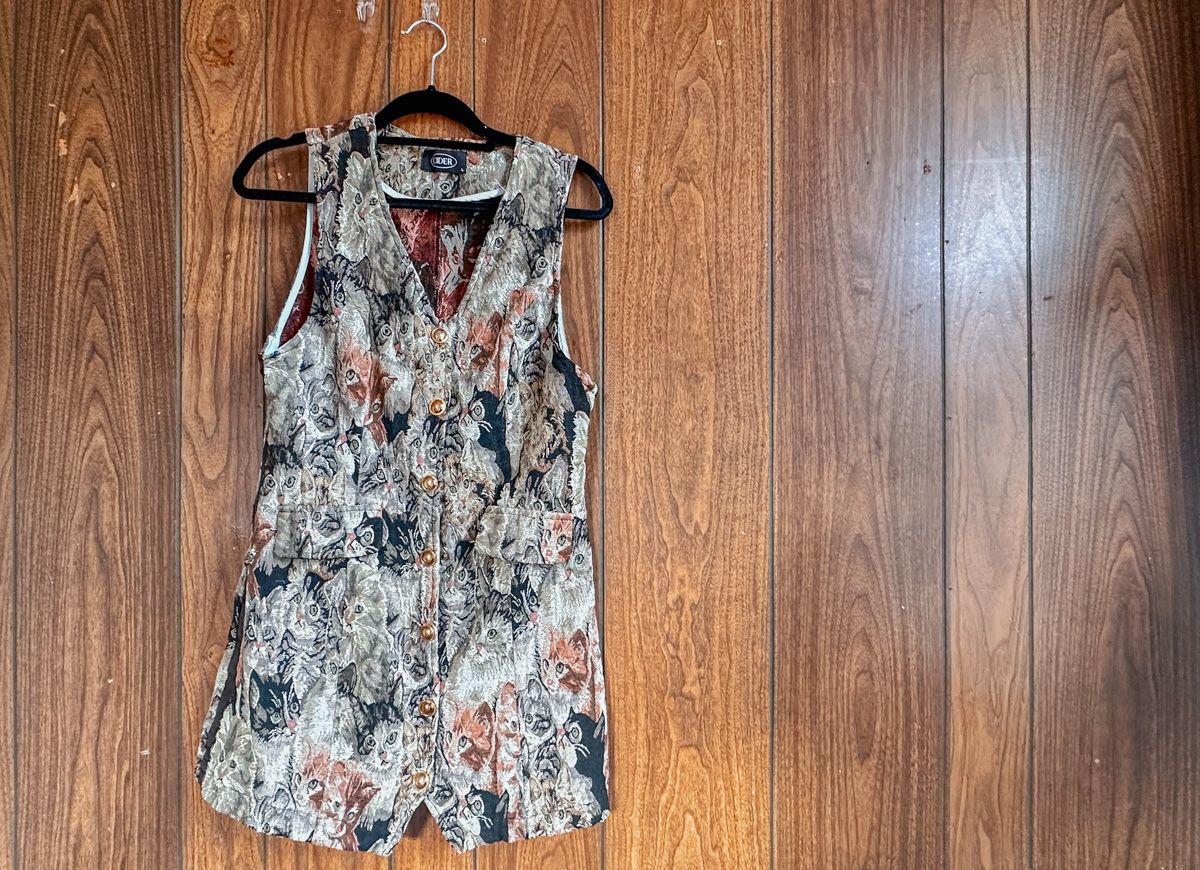 A mini-dress printed with earth-toned cats hangs on a hook against a wood-paneled wall.