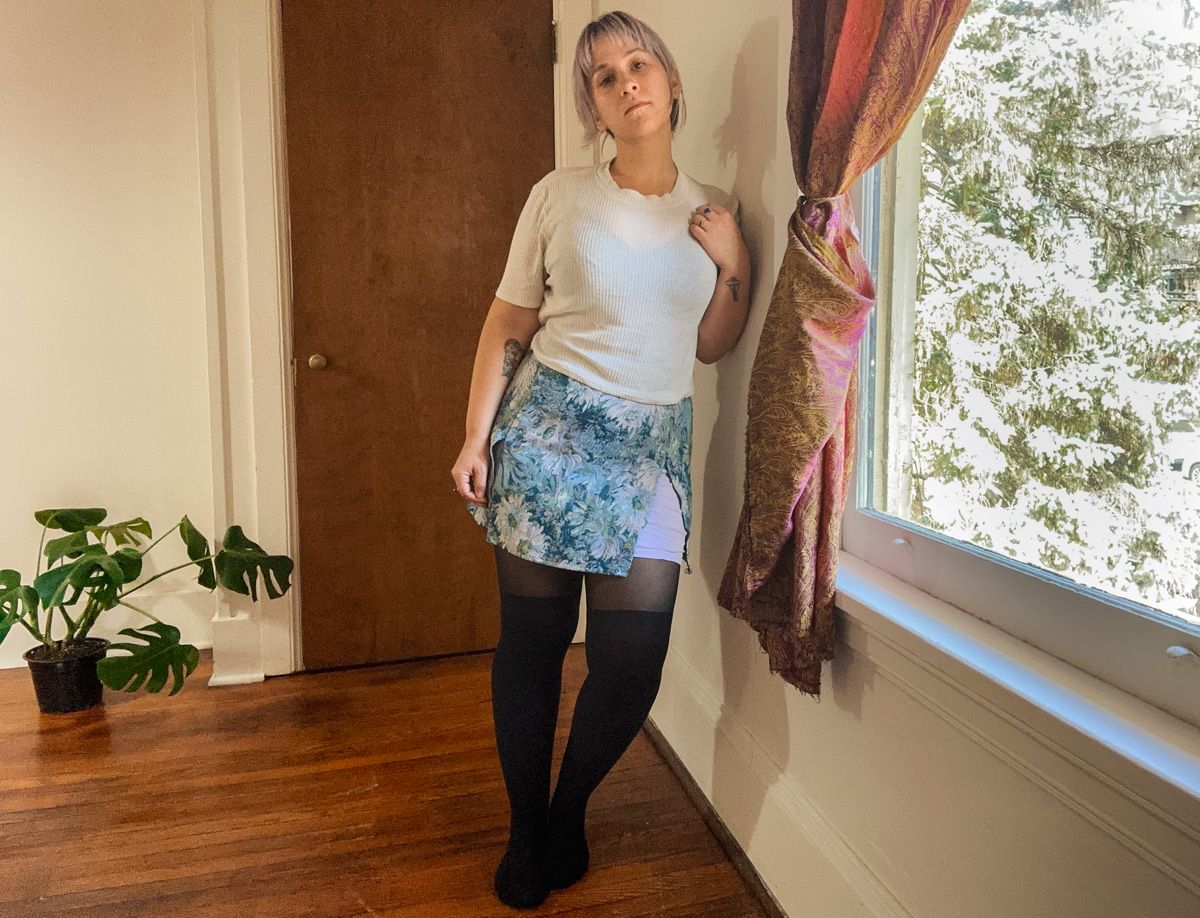 A purple-haired woman wearing a blue floral printed miniskirt and a white top stands facing the camera in a sparse interior space next to a window with trees visible outside.