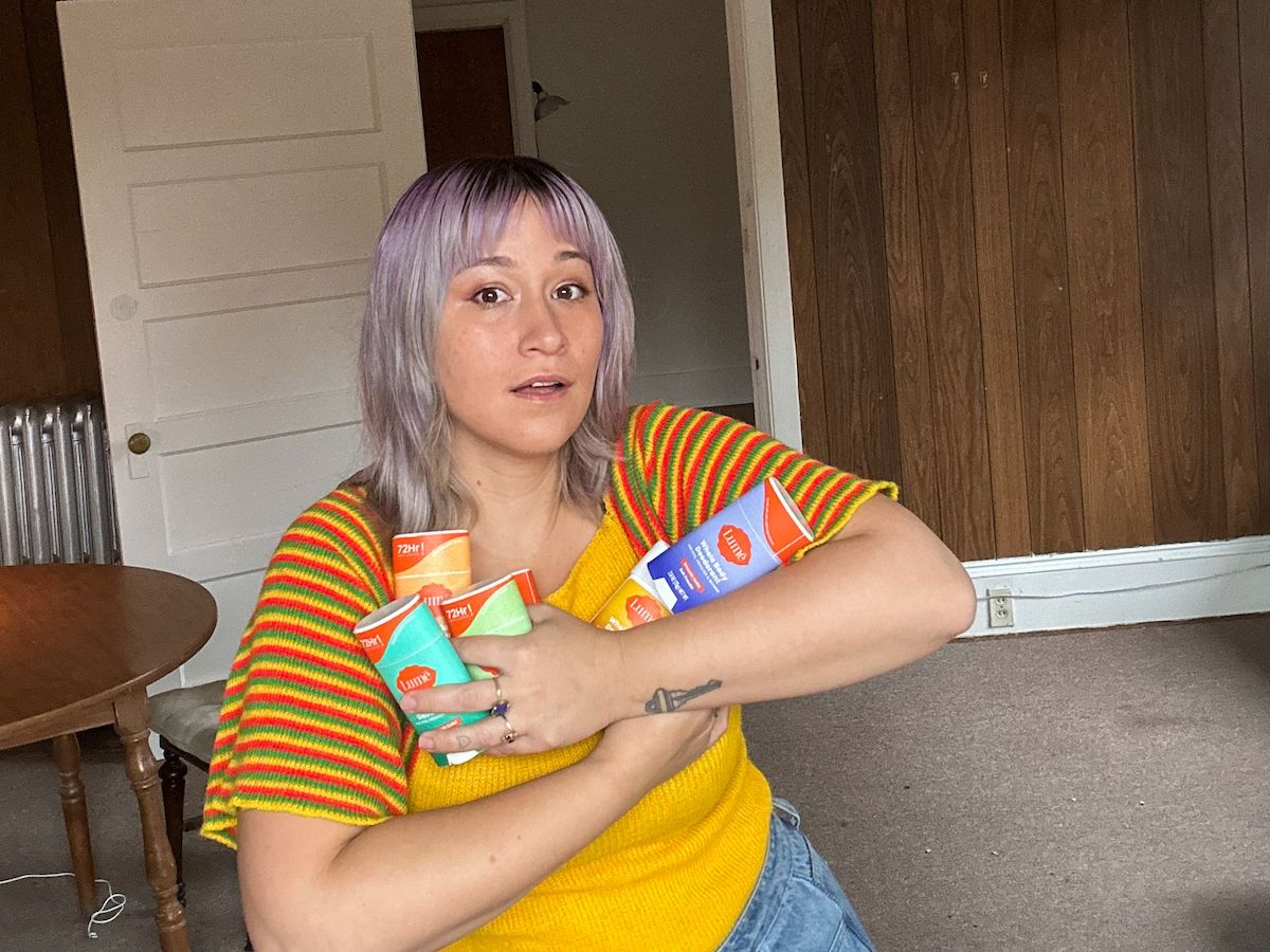 A woman with purple hair and a bright yellow shirt looks surprised as she attempts to juggle many sticks of deodorant, with a wood-paneled interior in the background.