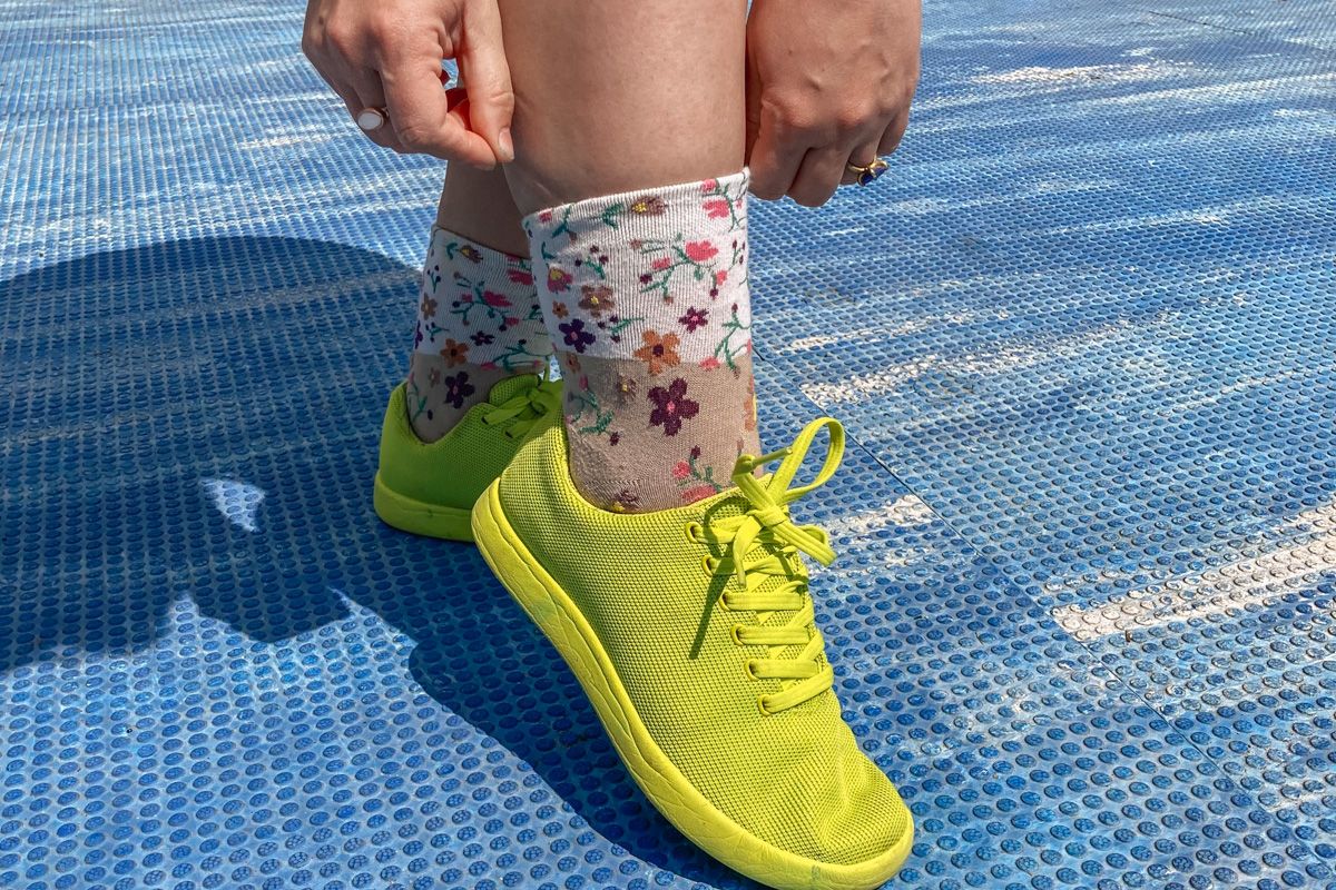 Close-up of a pair of feet wearing neon green shoes standing on a blue surface, with hands pulling up a pair of floral-print, white and beige socks.