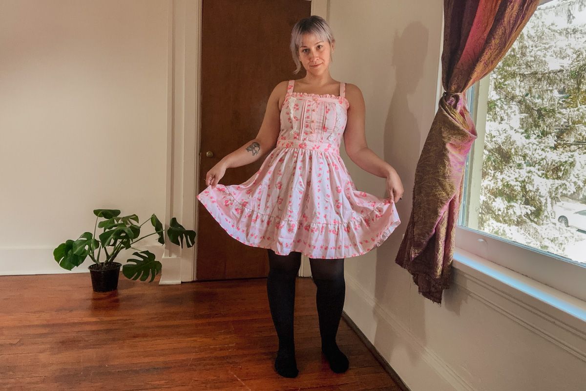 A purple-haired woman wearing a pink frilly dress lifts her skirt in a curtsy while standing in a sparse interior space beside a window where leafy branches are visible outside.