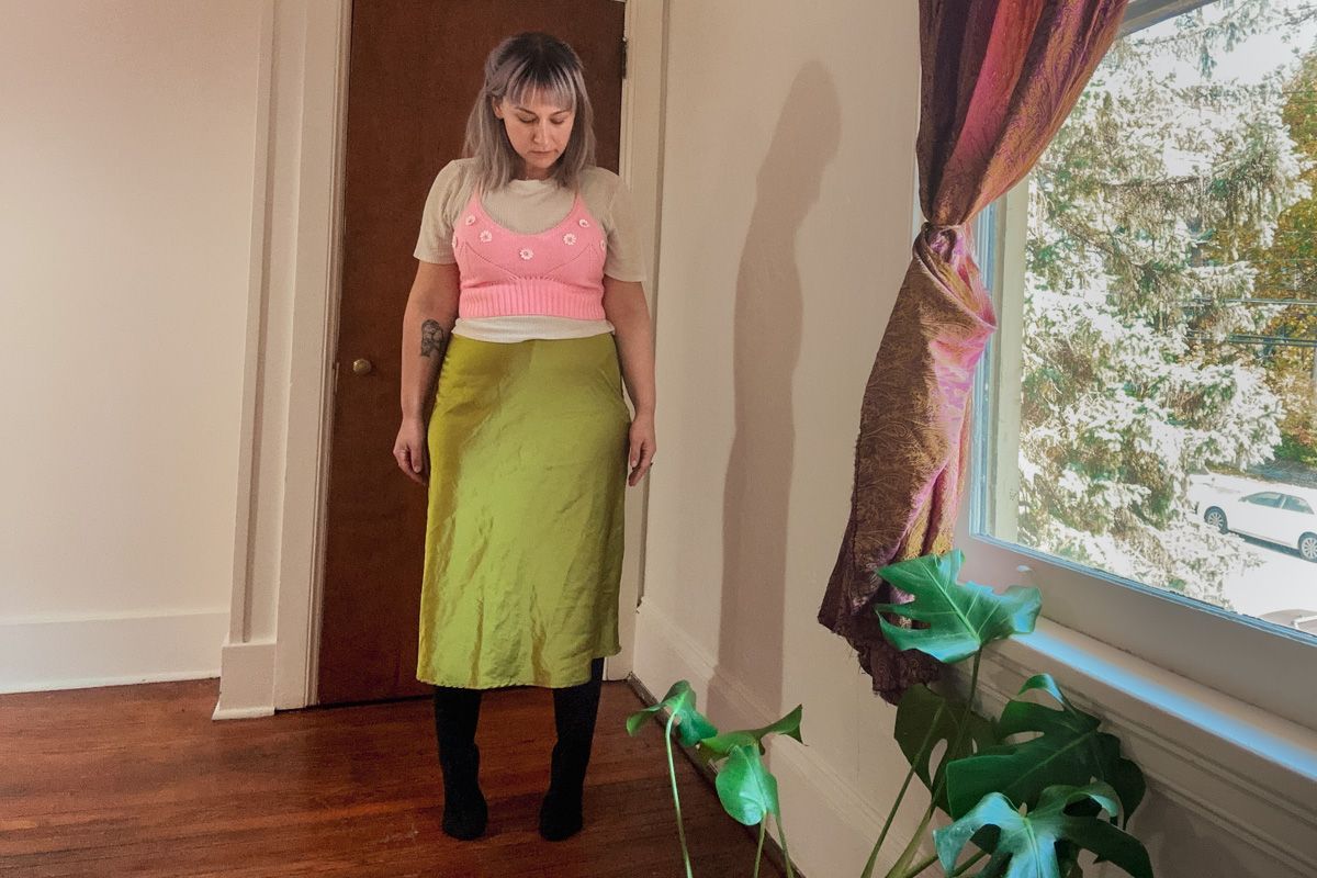 A purple-haired woman wearing a pink tank top and a long, silky, chartreuse skirt stands looking down in a sparse interior space beside a window where leafy branches are visible outside.