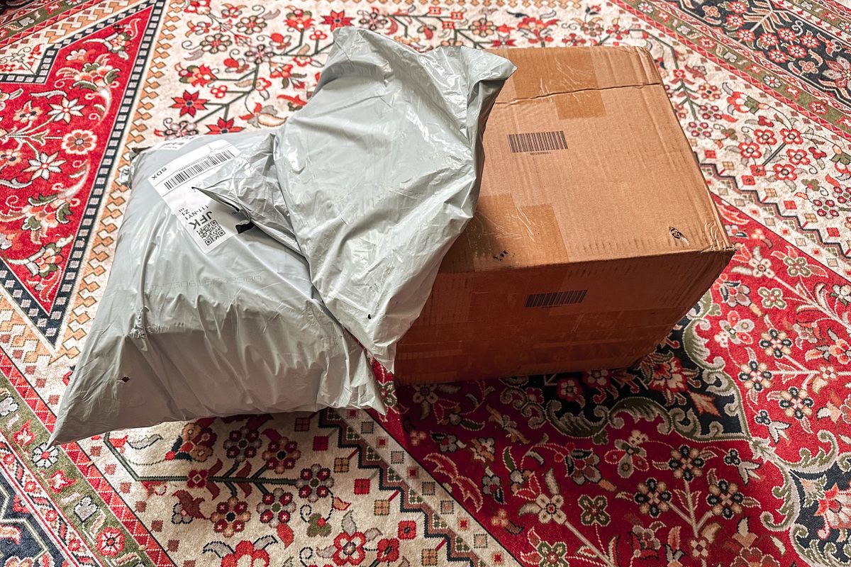 A cardboard box and several light grey soft postal packages sit on a red and white Persian rug.