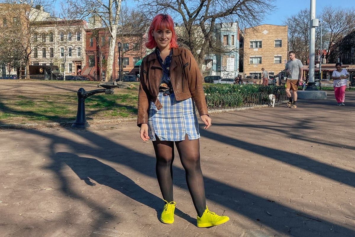 A woman with pink hair in a brown leather jacket, a blue plaid miniskirt, and neon green shoes smiles towards the camera in a city park.