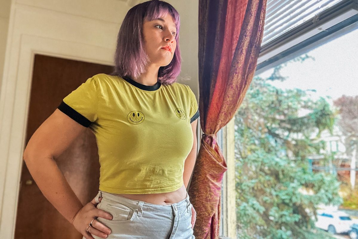 A woman with purple hair and a yellow t-shirt printed with a happy face and a sad face stands with with her hands on her hip, a sparse interior and a window overlooking trees behind her.