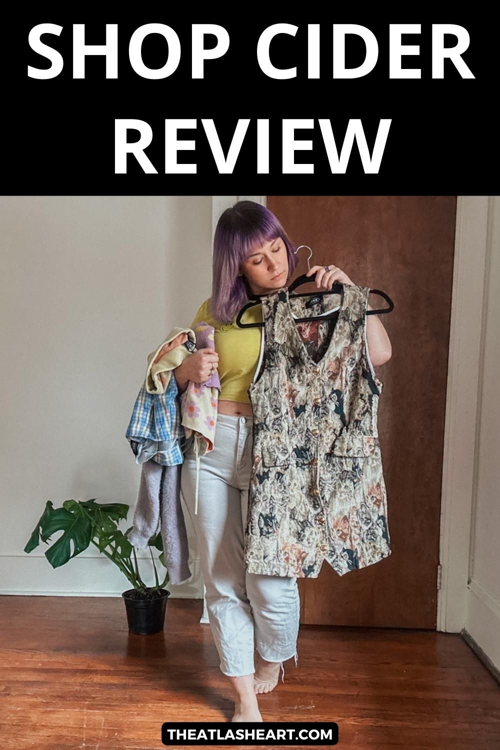 A sparse apartment interior with a woman with purple hair wearing a yellow shirt holding a stack of clothing and a brown dress on a hanger up to her body with the text overlay, "Shop Cider Review."