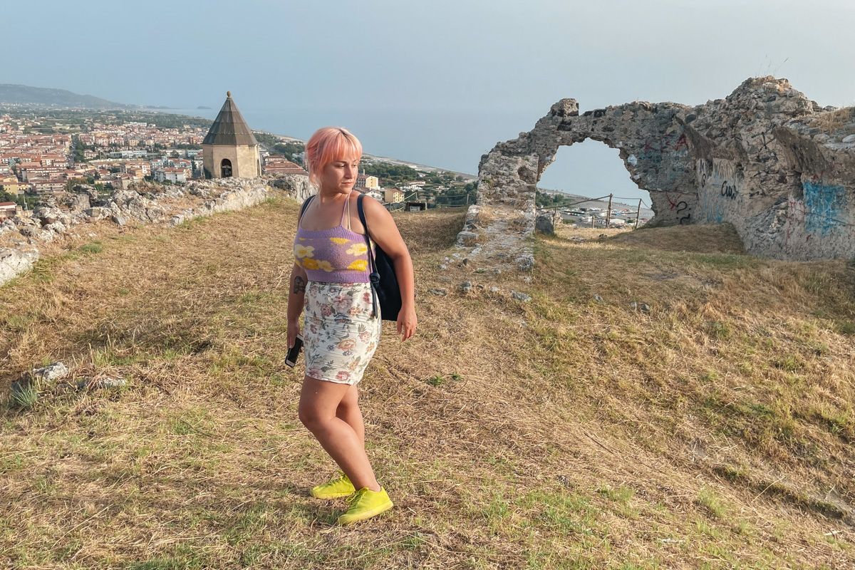 A woman with pink hair wearing a purple flower-print knit crop top, a white floral print mini-skirt, and neon green shoes stands in front of a crumbling stone doorway of an ancient ruin on top of a grassy hill.