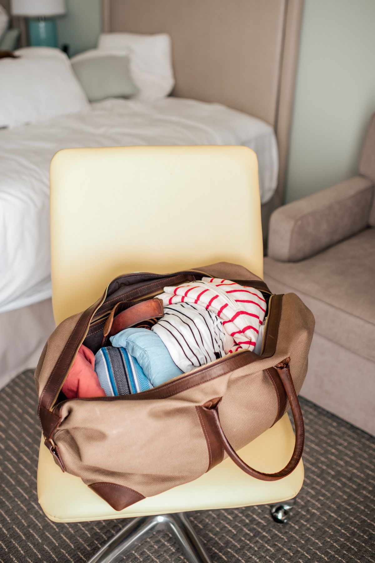 A brown duffel bag, open and filled with folded clothes, sits on a cream-colored chair in a bedroom suggesting packing for a trip.