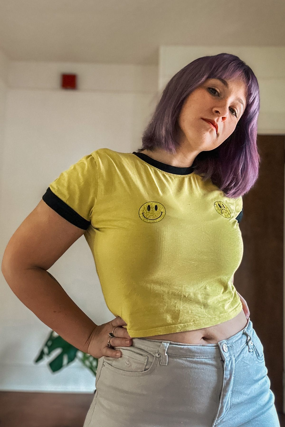 A woman with purple hair and a yellow t-shirt printed with a happy face and a sad face cocks her head to the side with a hand on her hip, a sparse interior in soft focus behind her.