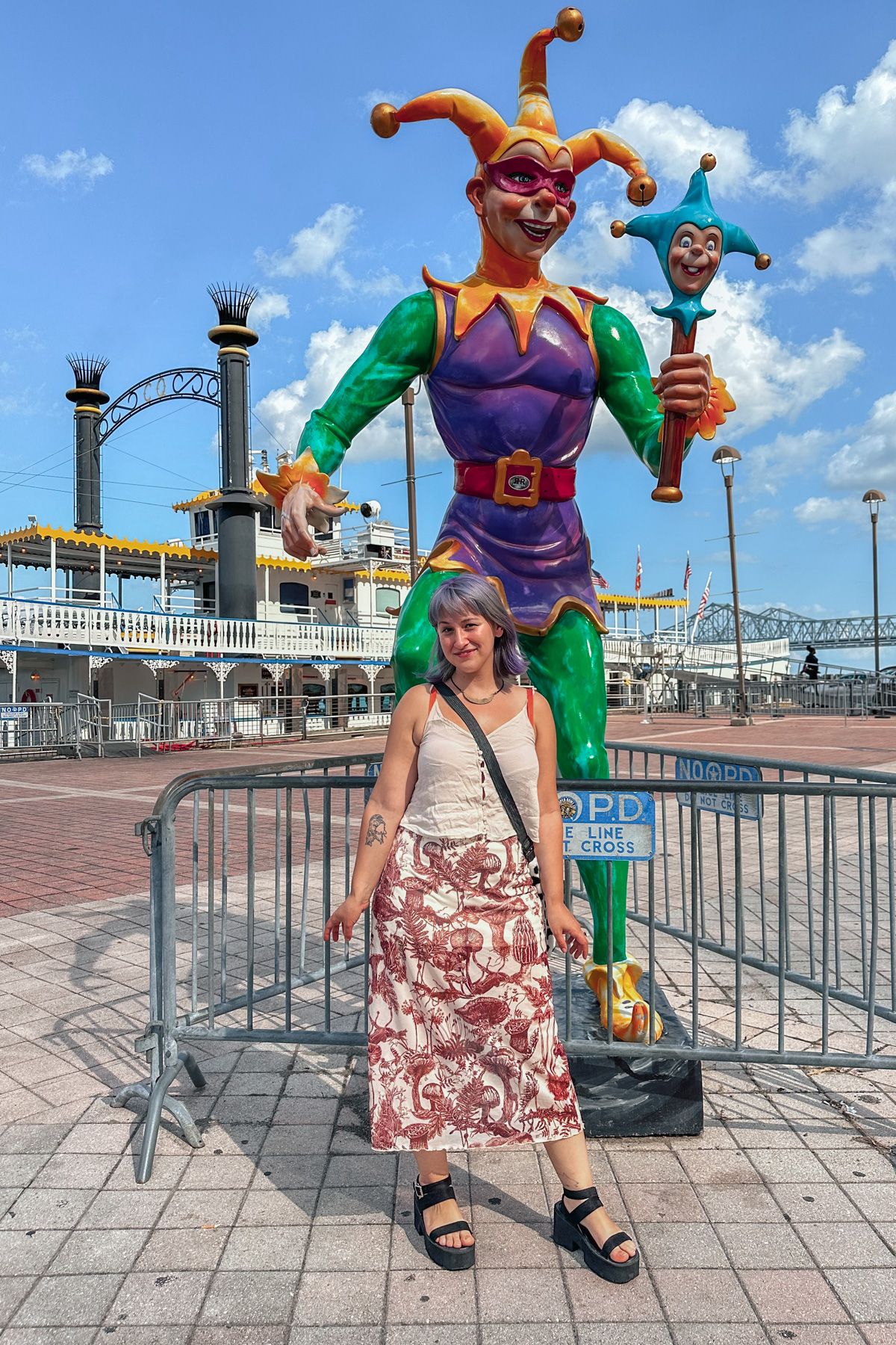 A purple-haired woman wearing a long, mushroom-printed skirt and a white tank top smiles as she stands in front of a statue of a purple and green jester, with a riverboat in the background.
