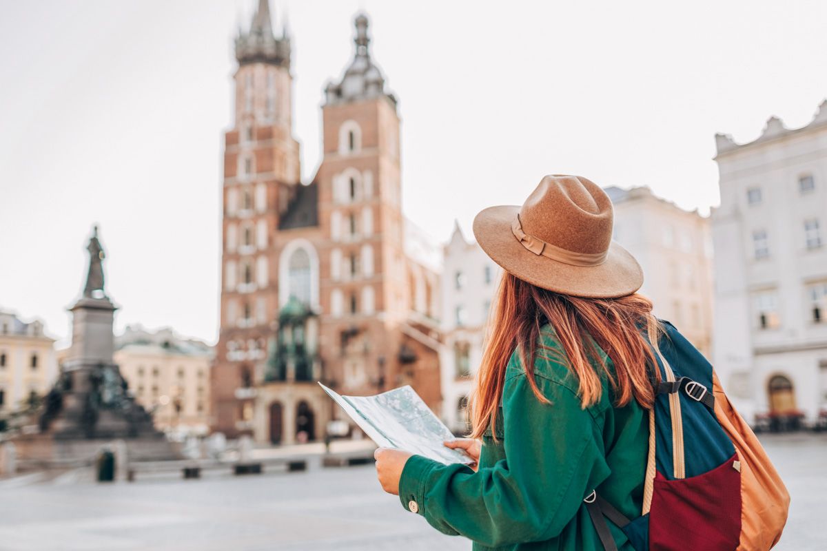 A female traveler seen from behind wearing a brown, wide-brimmed hat and a green jacket holds up a map she she stands in a European town square. 