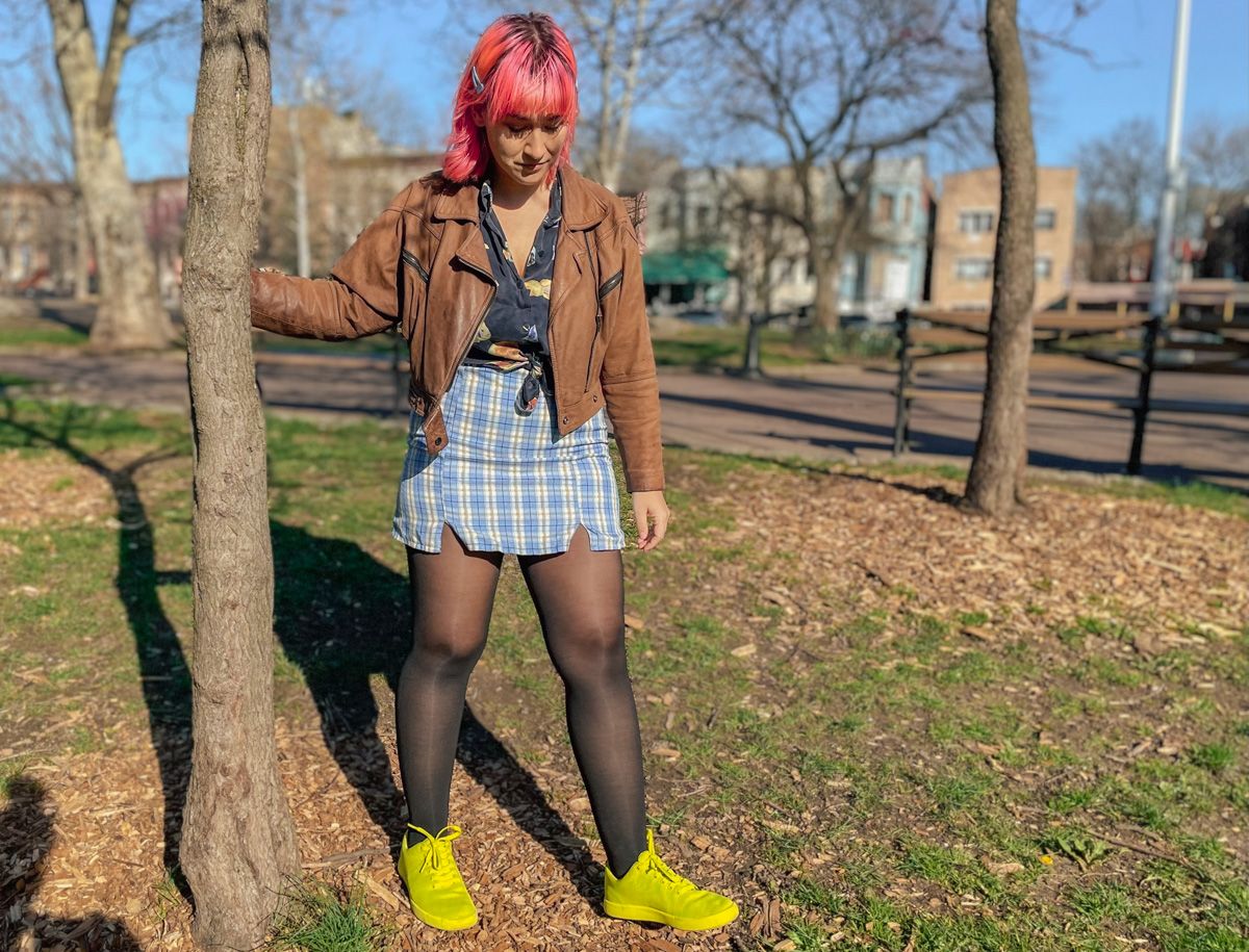 A woman with pink hair in a brown leather jacket, a blue plaid miniskirt, and neon green shoes stands next to a tree in a city park.