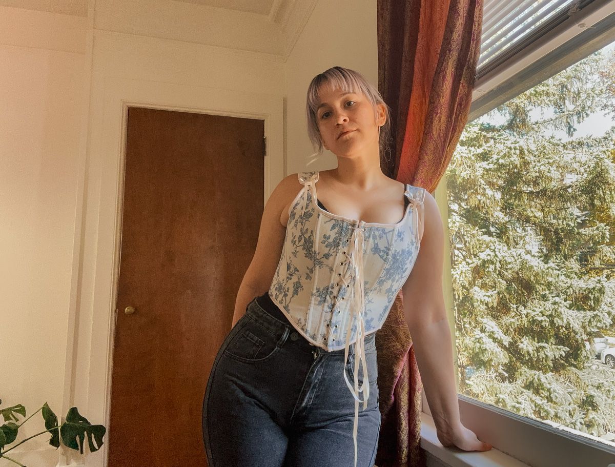 A purple-haired woman wearing a blue-and-white toile corset top stands with a hand on the windowsill in a sparse interior space beside a window overlooking leafy branches.