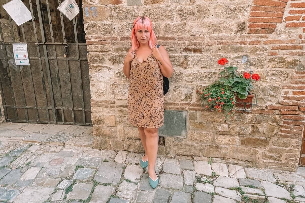 A woman with pink hair and a cheetah-print dress wearing a pair of teal Vivaia flats and standing against a wall on a cobblestone street.