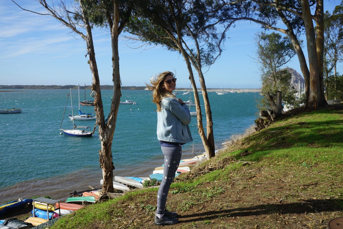 A woman wearing navy blue patterned leggings and a jean jacket smiles at the camera while standing on a grassy embankment with a view of a sunny harbor framed by eucalyptus trees visible behind her.