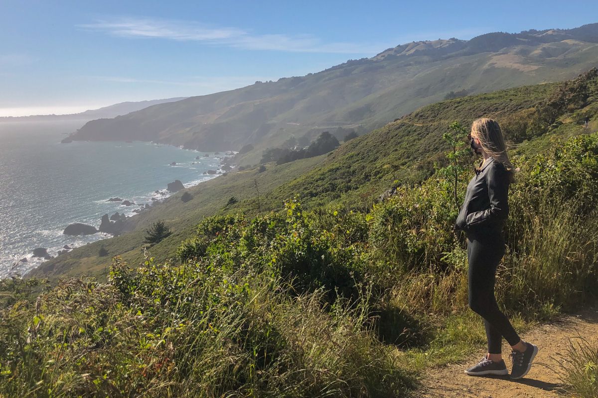 A woman wearing black leggings and a leather jacket looks out at the view while standing on a grassy hill looking out over an ocean view.