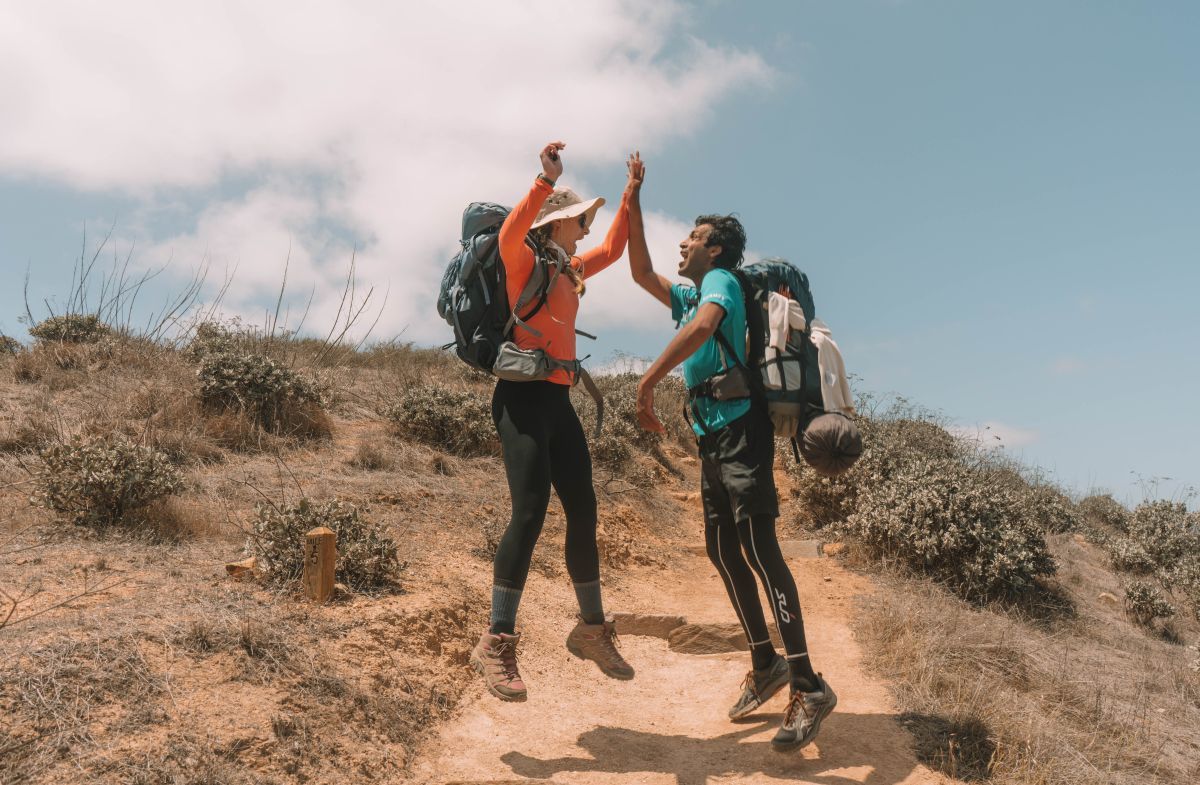 A man and a woman in hikinggear and hiking backpacks jump in the air and high five on a rocky path in an arid landscape.