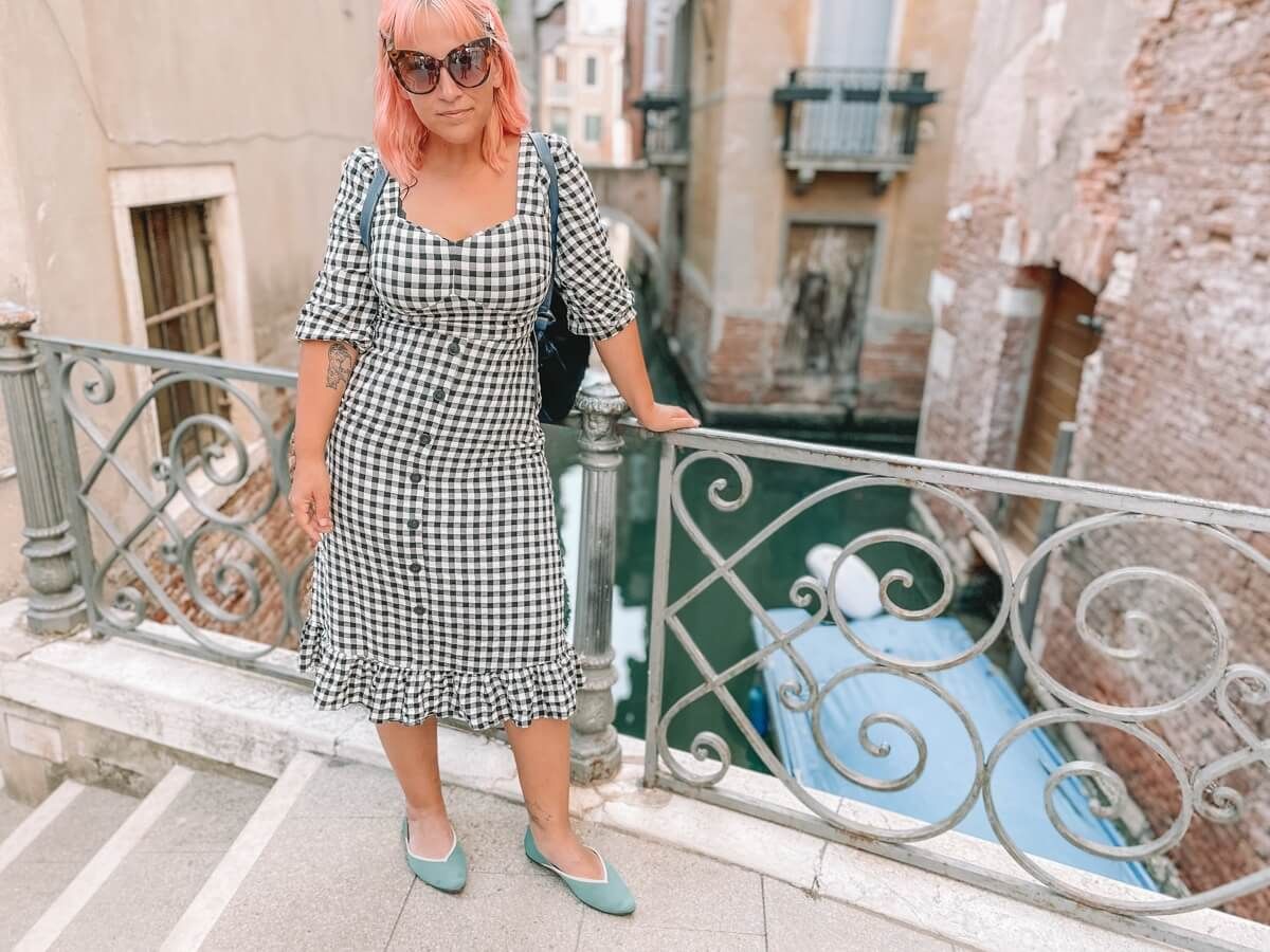 A woman with pink hair, sunglasses, and a black-and-white checkered dress wears A pair of teal Vivaia flats on a bridge over a Venetian canal.