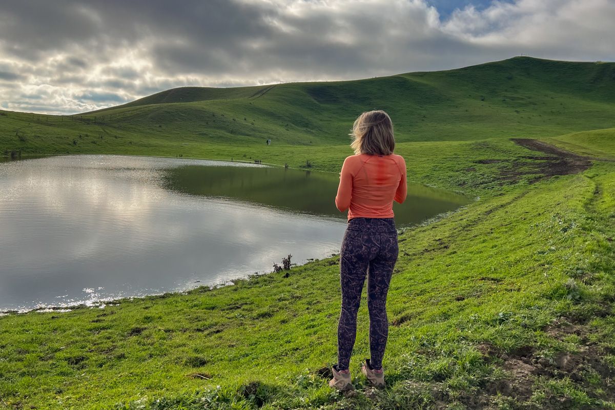 A woman in a pink shirt and patterned leggings stands with her back to the camera in a grassy, hill, landscape looking at a small, calm pond.