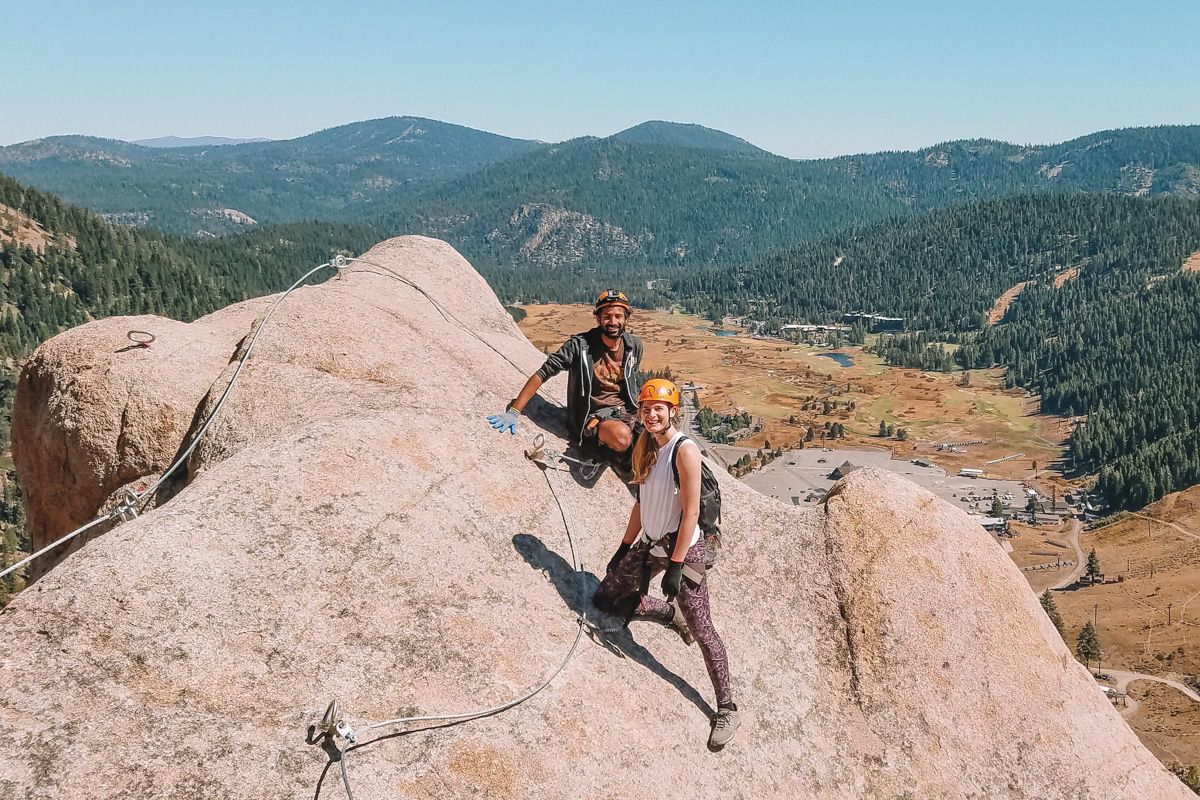 A man and woman wearing activewear and rock-climbing gear smile at the camera while standing on a boulder overlooking an alpine valley. 