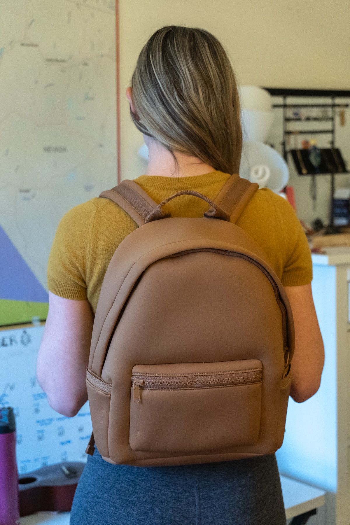 A young woman wearing a brown Neoprene Backpack, grey Ultra-Soft High-Rise Leggings and a yellow shirt stands with her back to the camera in an interior setting.