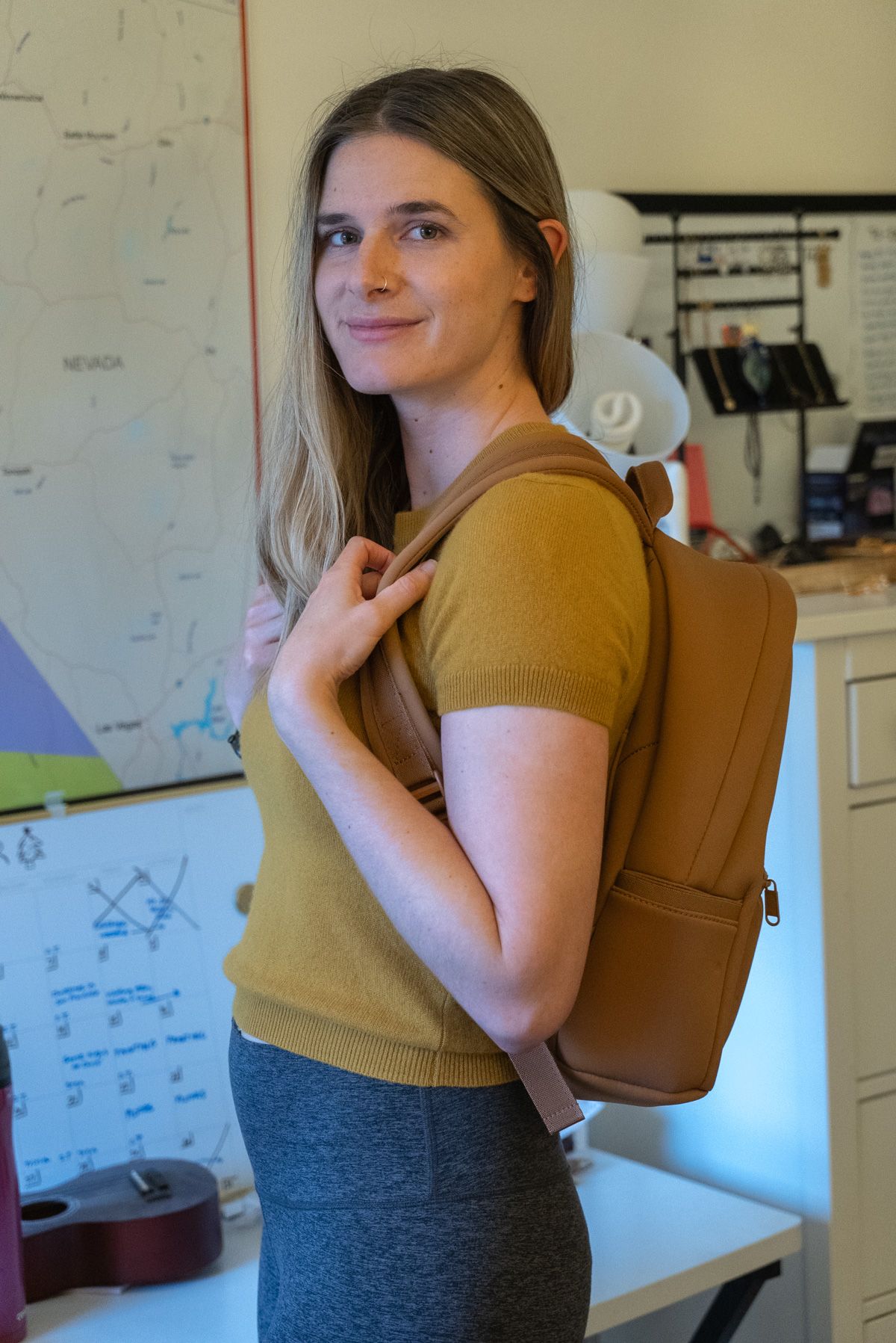A young woman wearing a brown Neoprene Backpack, grey Ultra-Soft High-Rise Leggings and a yellow shirt stands to the side looking over her shoulder at the camera in an interior setting.