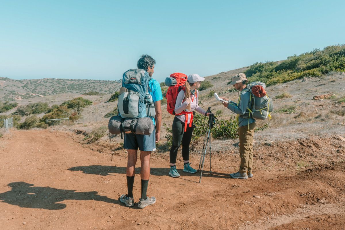 A male hiker wearing tall hiking socks and shorts stands facing a male and female hiker who are consulting a map at the intersection of a red dirt road in a dry landscape.