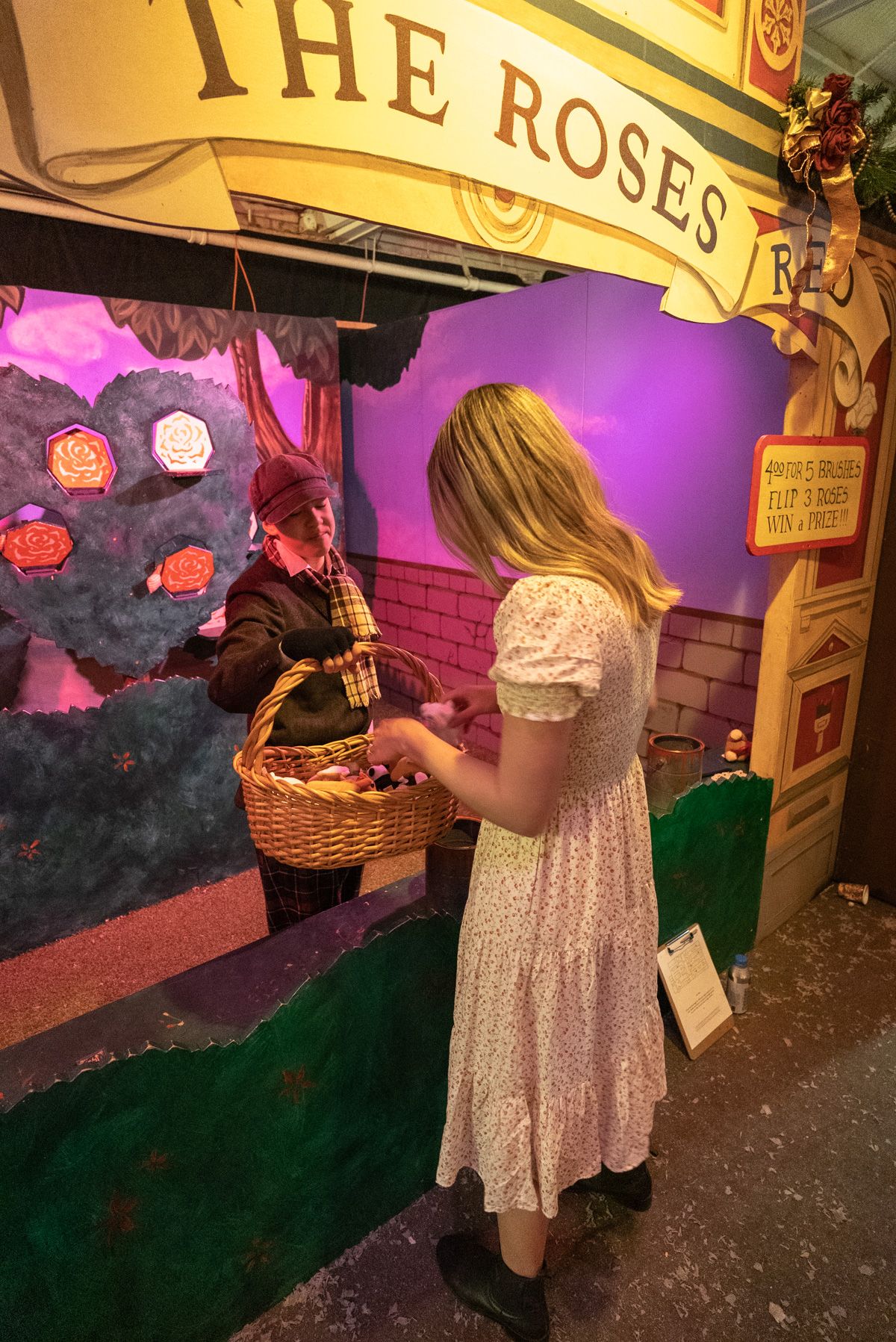 A woman in a white dress seen from behind at a Victorian-style Carnival Games booth with a sign that reads, "The Rose."