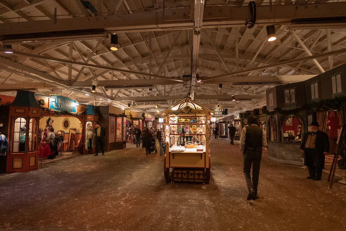 People in Victorian costumes mill around an indoor Christmas market designed to look like a shopping street in Victorian London.