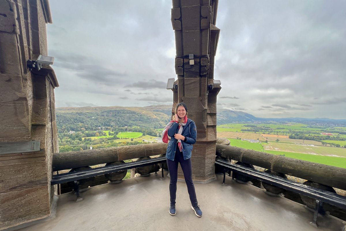 A young woman in black pants and a dark blue jacket stands on a stone balcony in front of a view of rolling countryside hills and an overcast sky.