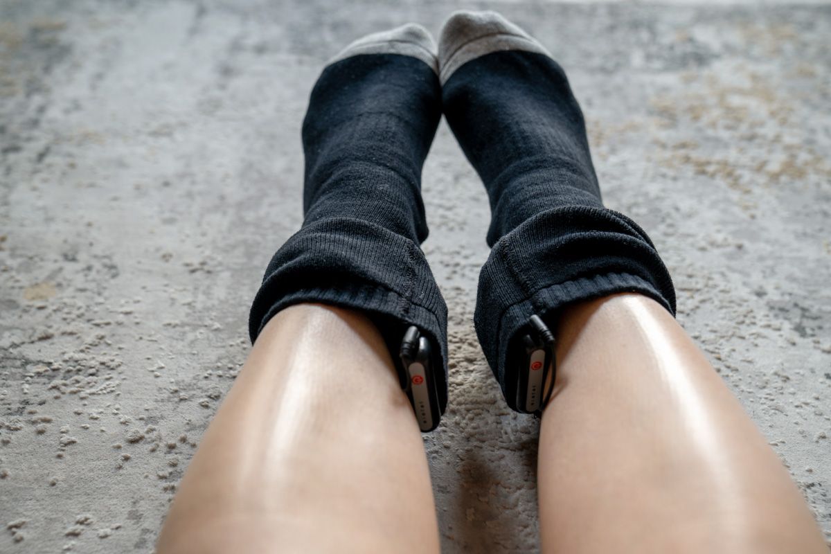 Point of view of a pair of legs and feet wearing black heated ski socks with battery packs, scrunched down to the ankles, sitting against a textured grey surface.