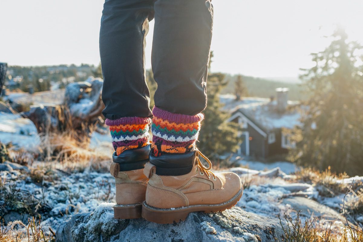 A pair of feet in tan boots and thick multi-colored socks, seen from behind standing on a frost-dusted rock looking out at a view.