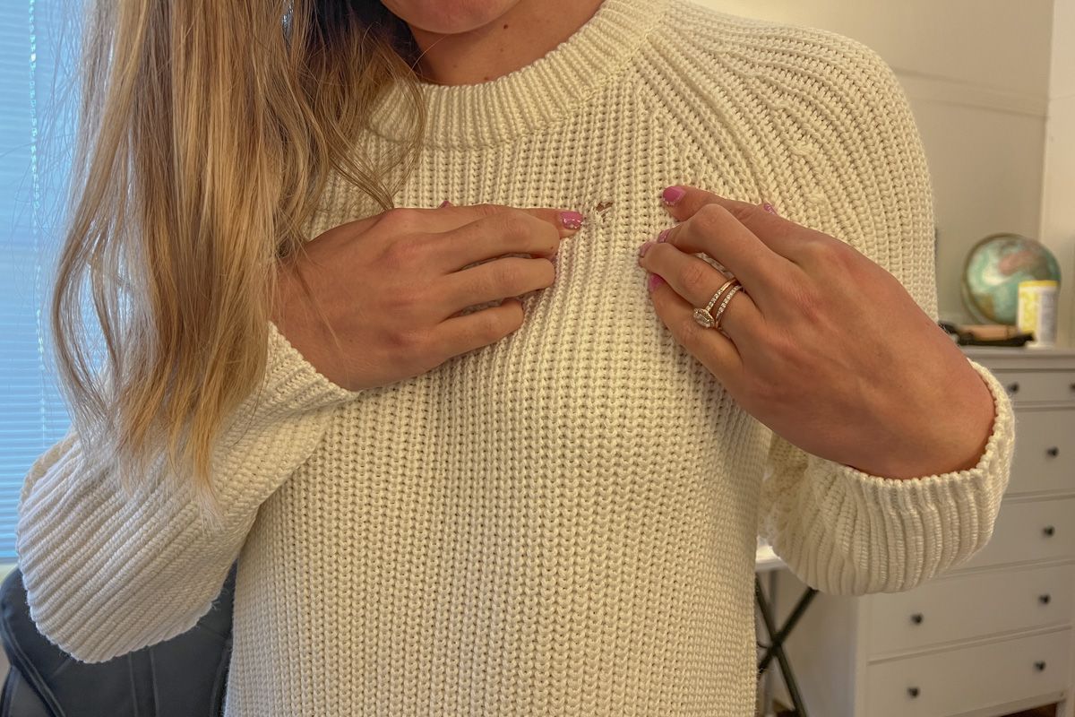 A woman wearing a white sweater seen from the shoulders down, pointing to a small hole in the sweater's chest.
