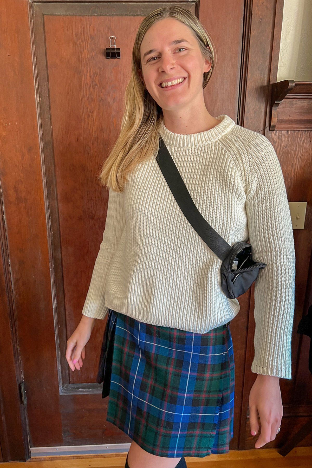 A young woman wearing a white sweater and a blue plaid skirt stands smiles at the camera in front of a hardwood interior.