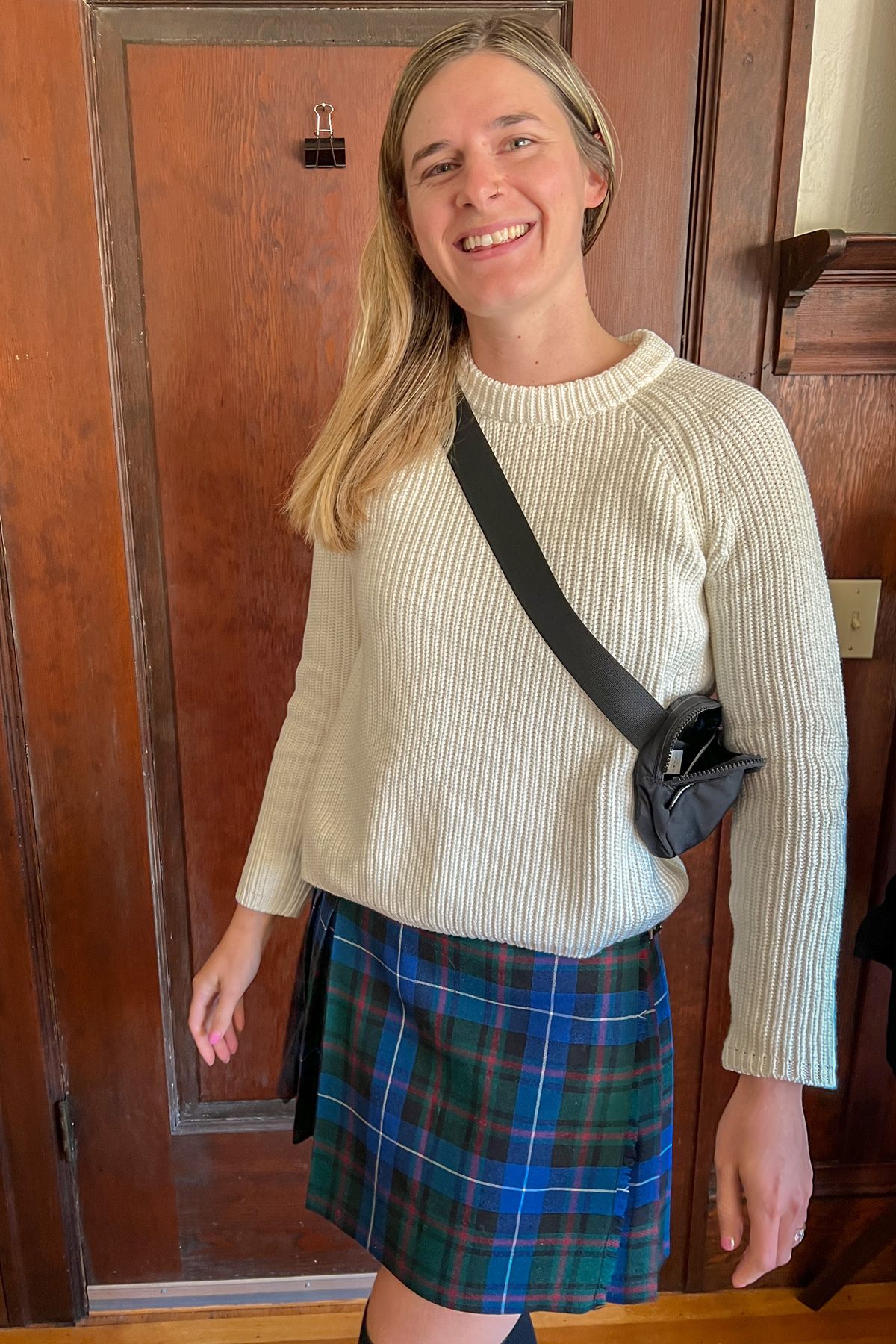 A young woman wearing a white sweater and a blue plaid skirt stands smiles at the camera in front of a hardwood interior.