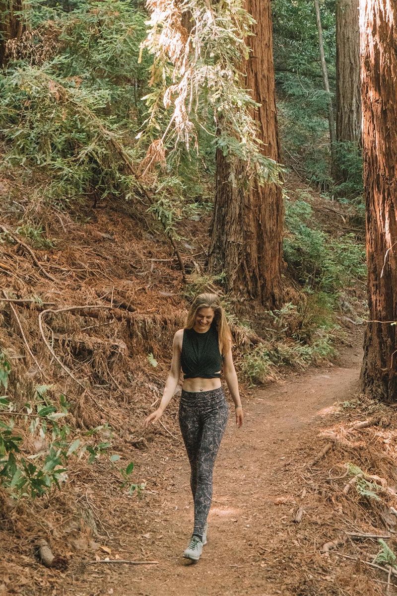 A woman wearing navy blue patterned leggings and a black tank top looks down while walking towards the camera down a path through a redwood forest.