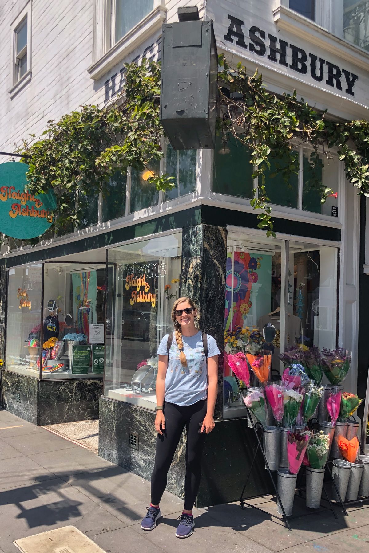 A woman in black leggings and a blue shirt stands smiling on a street corner in front of a store with a sign that says "Haight Ashbury."
