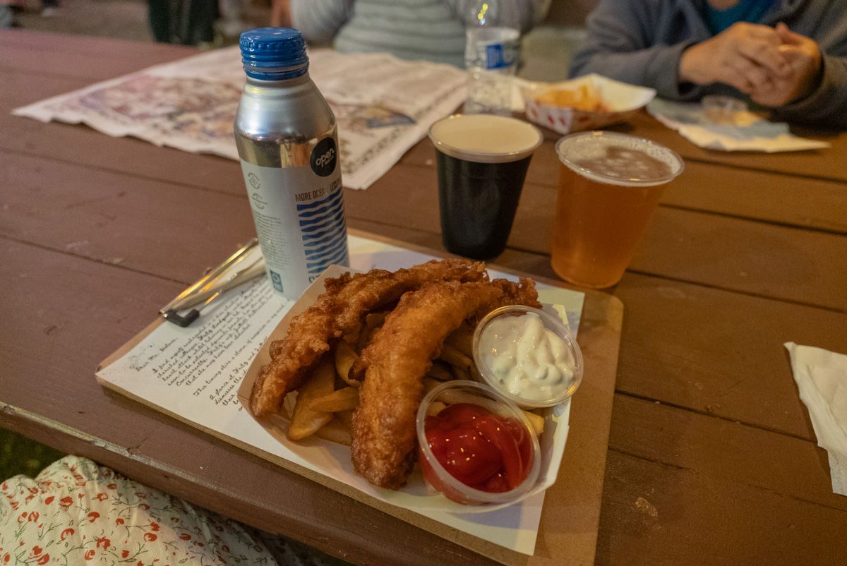 A close-up of a fish and chips meal with two beers in plastic pint glasses sitting on a brown wood picnic table.
