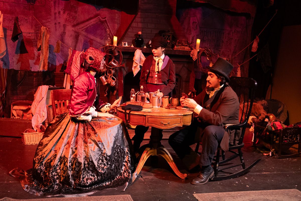 Two performers in Victorian costumes sit across each other at a table on a stage, lit with red light, with a small boy standing upstage behind them.
