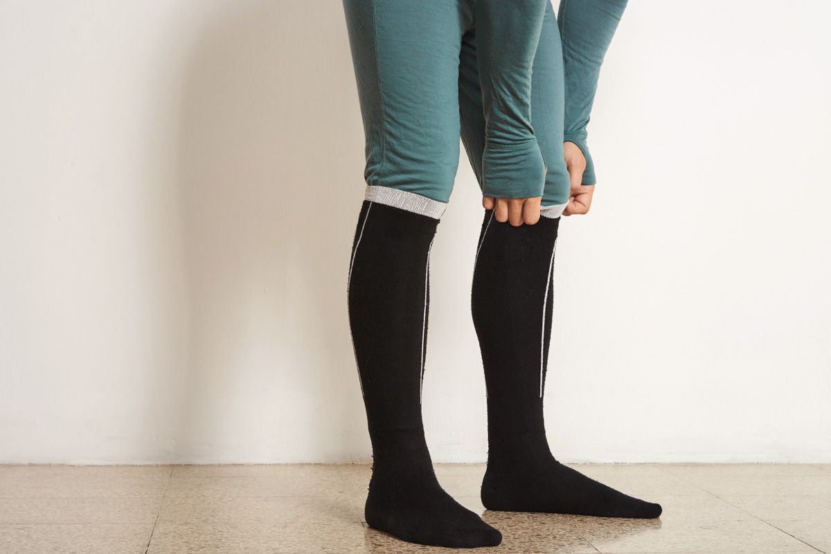 A pair of legs and feet wearing black heated ski socks and blue long underwear standing in front of a blank white wall.
