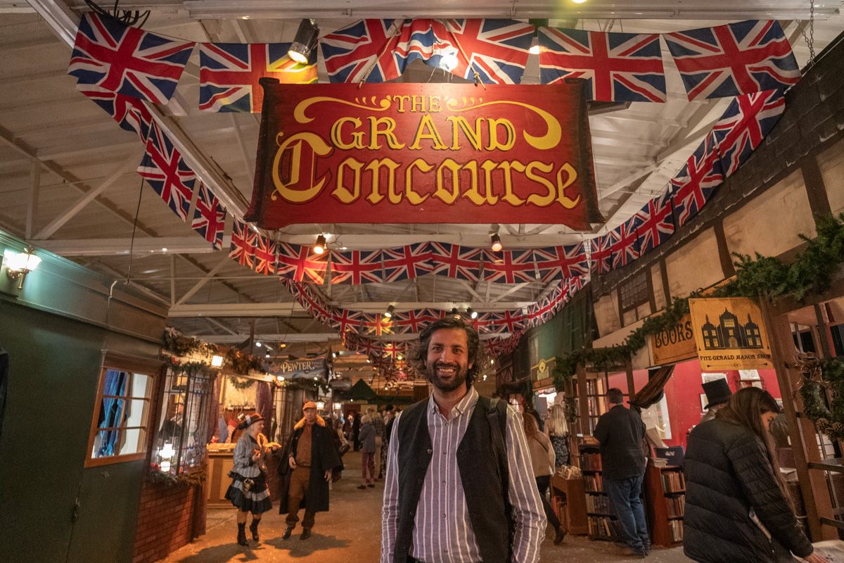 A man in a pin-striped shirt and brown vest smiles while standing in front of the red and gold Grand Concourse sign at the Dickens Fair in San Francisco