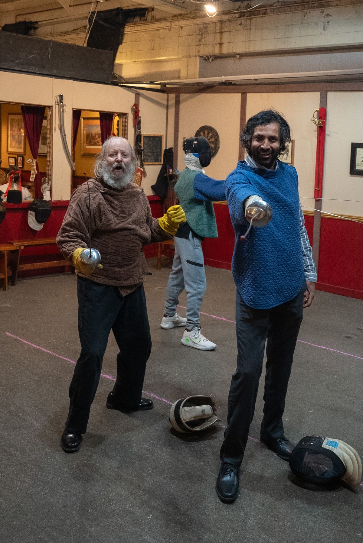 A man in a blue fencing outfit smiles beside an older man with a white beard as they hold up swords in a fencing studio.