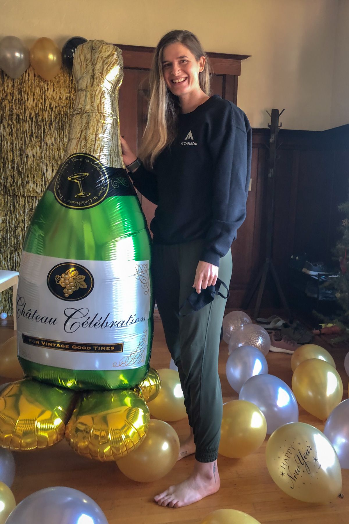 A woman wearing green joggers and a dark sweatshirt smiles at the camera in a living room, standing next to a giant balloon shaped like a champagne bottle.