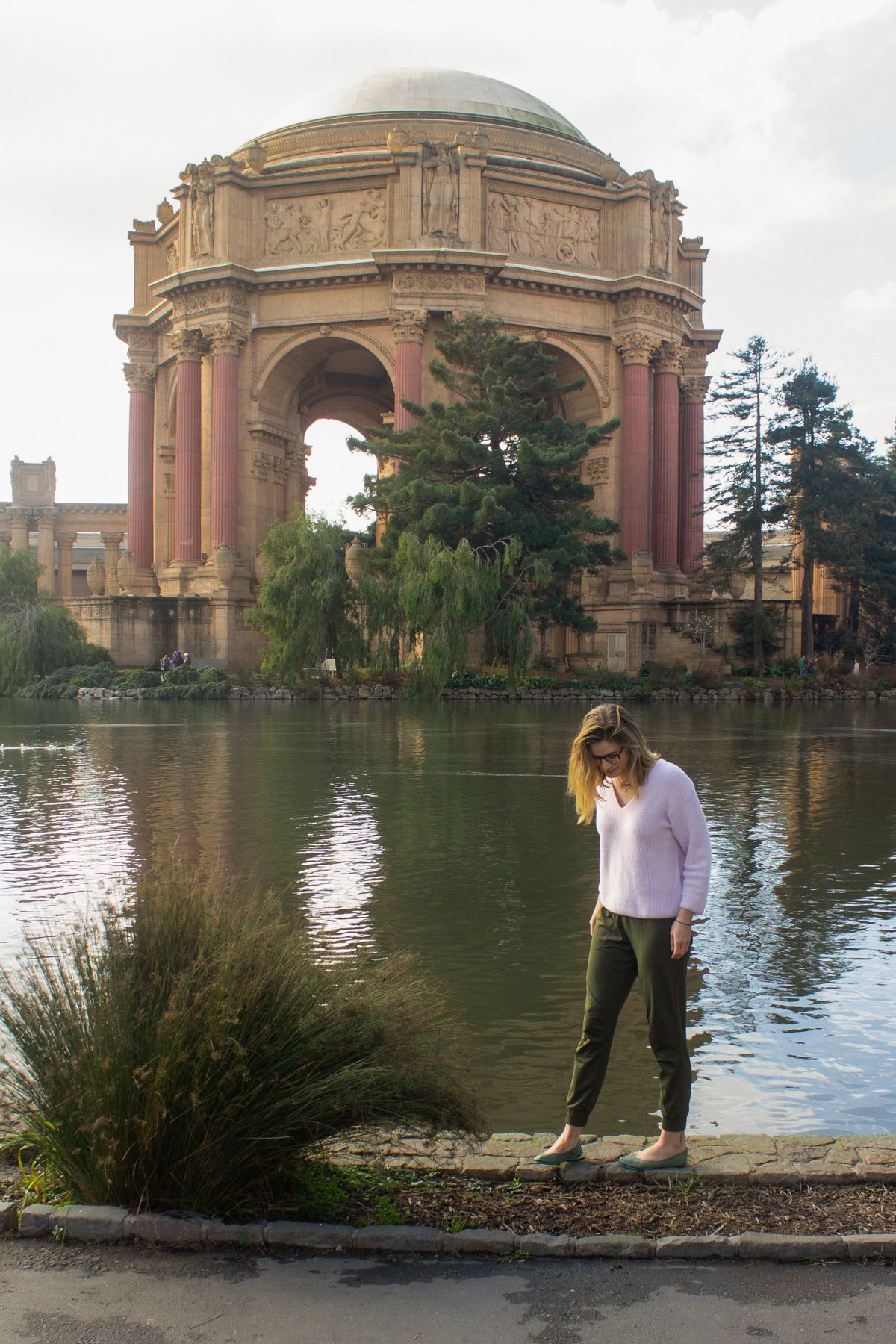 A woman wearing green joggers and a white sweater stands on the edge of a pond looking down at her feet with the Palace of Fine Arts behind her.